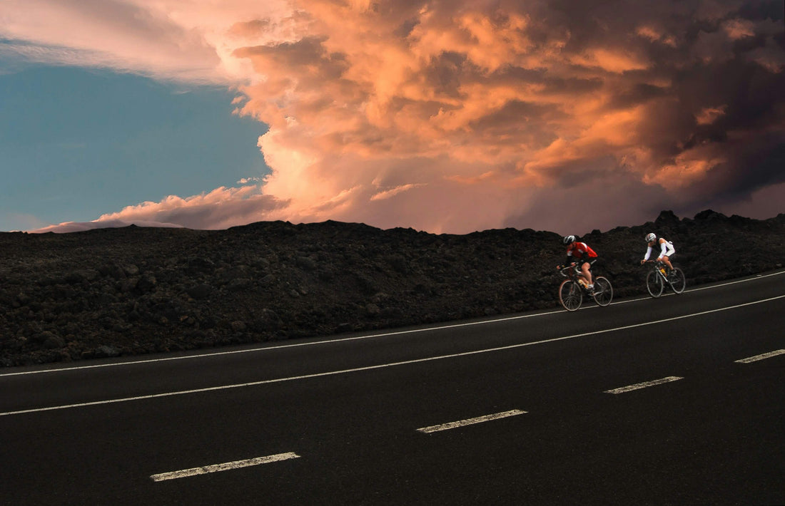 Two cyclists riding on a remote asphalt road with dramatic sunset clouds, demonstrating the importance of carrying a mini bike pump for long-distance adventures.